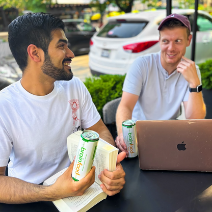 Two men sitting outdoors with a laptop, one holding brainfood sparkling water and a book.