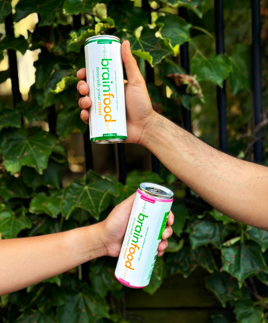 Two hands holding cans of Brainfood supplement in front of a green leafy background